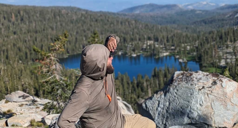 A person standing a high elevation shields their eyes from the sun. They are standing above an alpine lake framed by evergreens. 